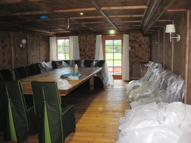 Stube mit dunkler Holzdecke und langem Tisch, die Stühle noch in Schutzfolie