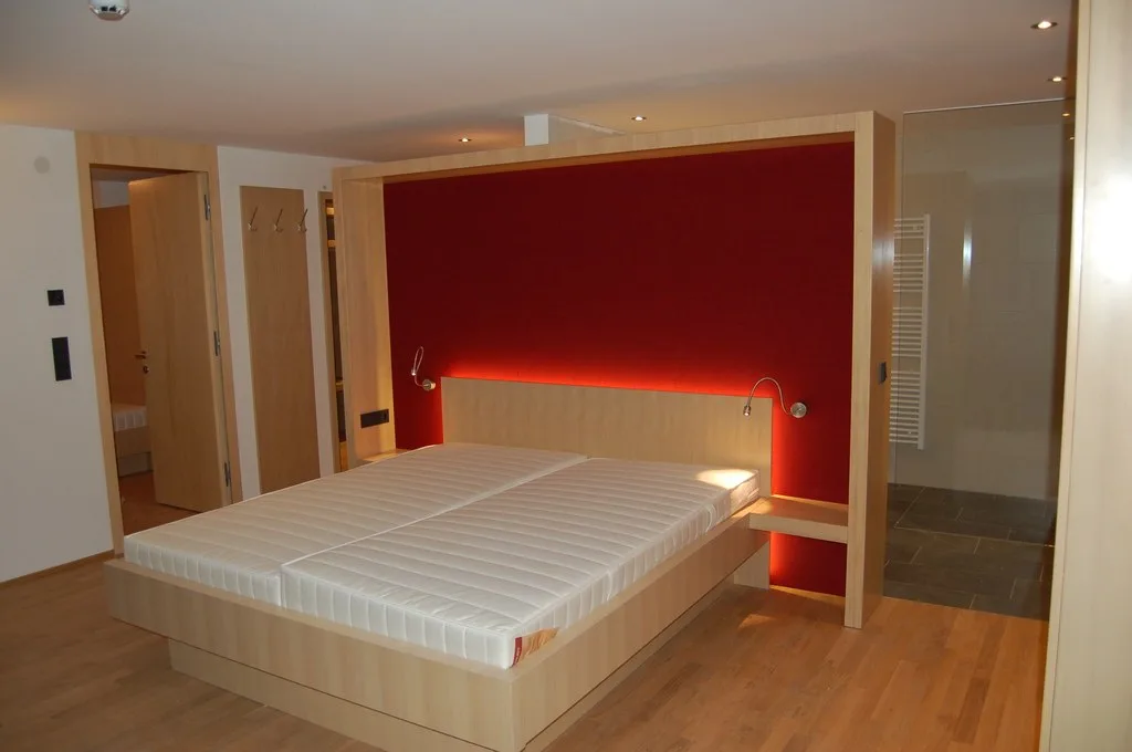 Hotelzimmer mit Bettrahmen und Wandscheibe aus hellem Holz vor einer roten, hinterleuchteten Rückwand