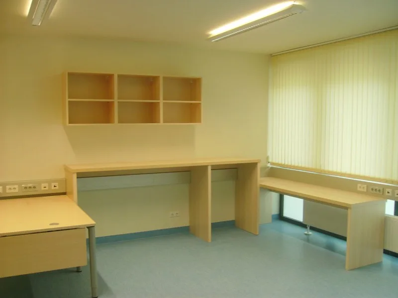 Raum mit hellen Schreibtischen in Holzoptik und einem offenen Wandregal