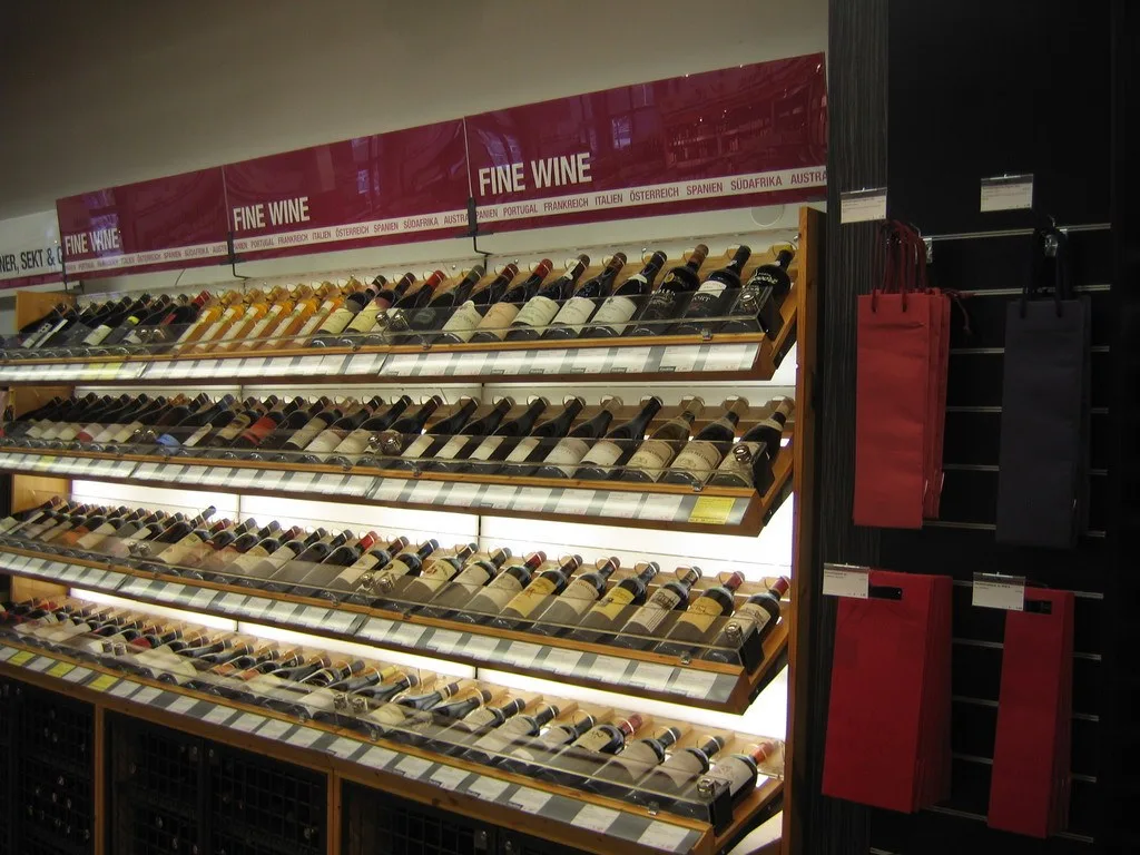 Beleuchtete Weinregale mit schrägen Fachböden und Schildern mit der Aufschrift Fine Wine, daneben rote Geschenktaschen