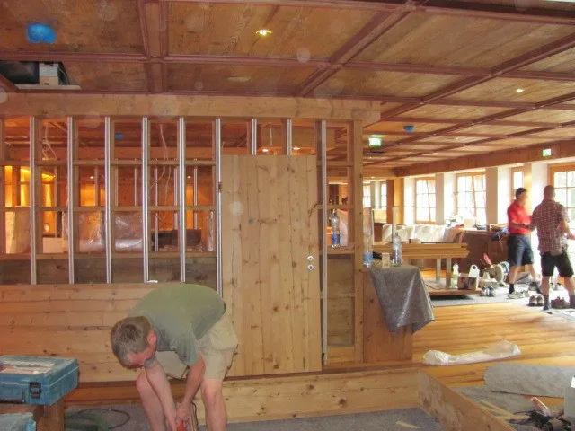 Monteure beim Ausbau einer Stube mit Holzdecke, einer kniet vorne bei einer Holzwand, zwei arbeiten im Hintergrund